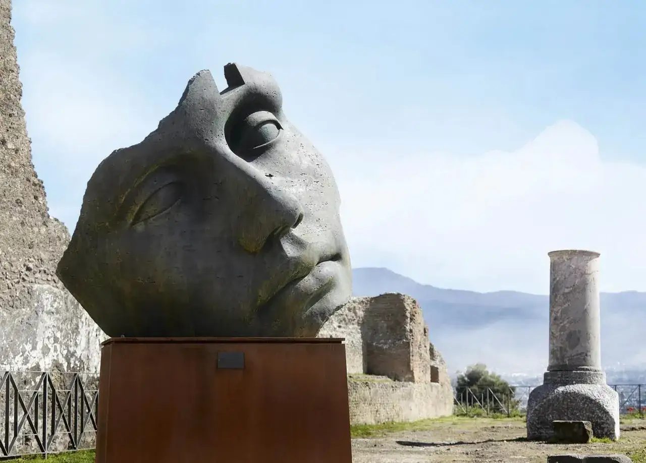 Kamienna rzeźba głowy z pęknięciami na tle ruin i gór.