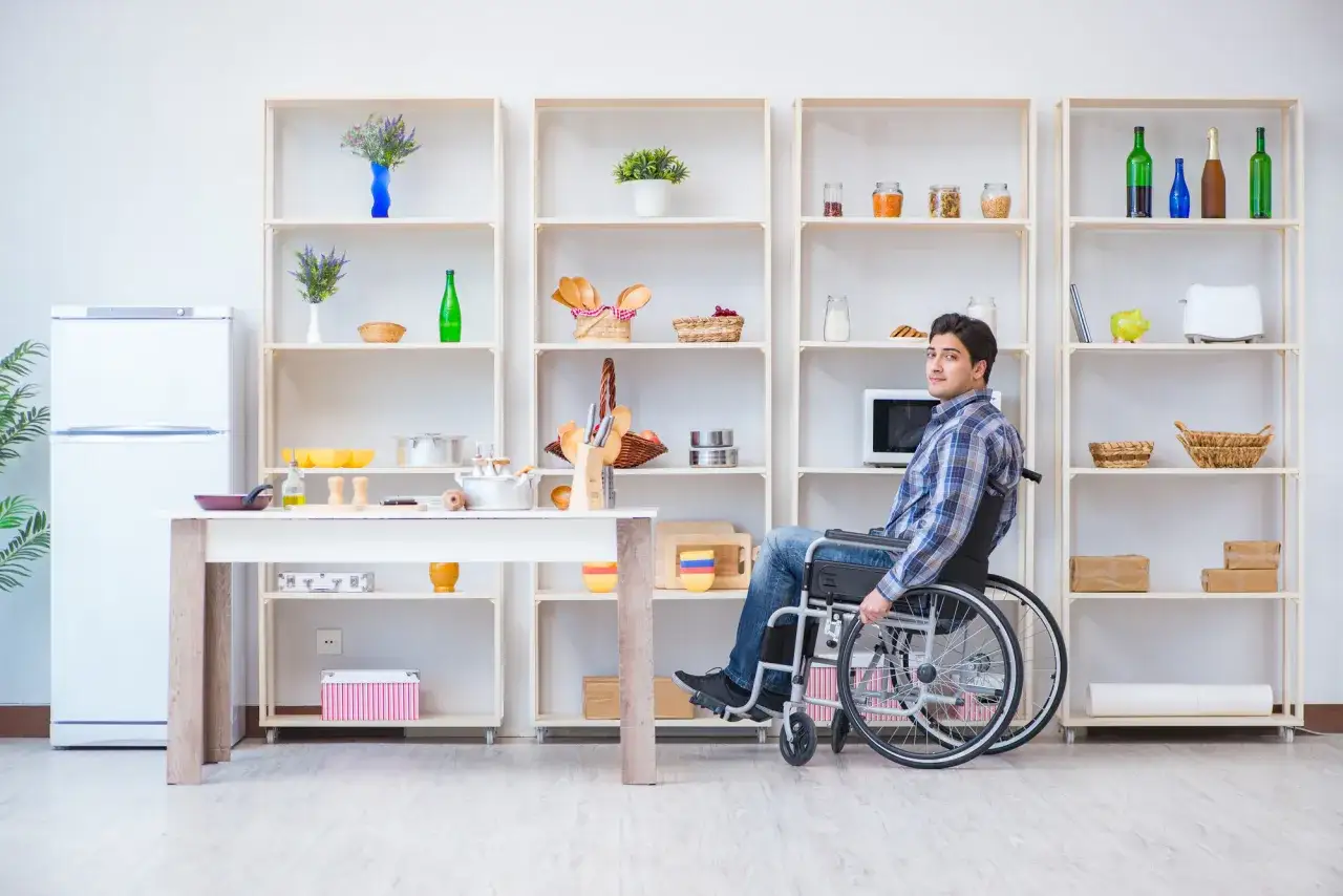 Mężczyzna na wózku inwalidzkim w kuchni, która jest przystosowana jako mieszkanie dla niepełnosprawnych. Wygodny dostęp do półek i stołu.
