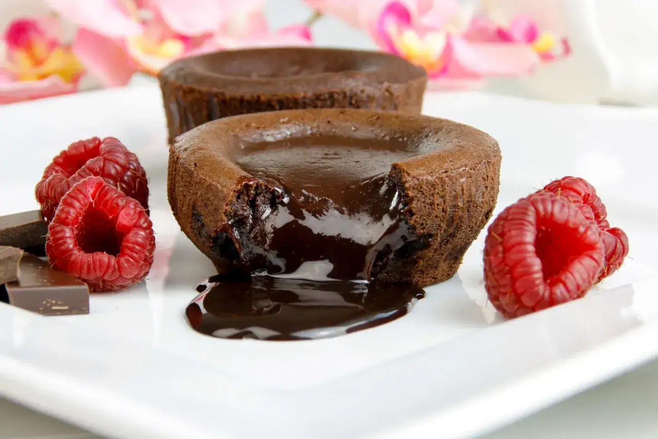 Płynny środek rozpływa się z gorącego fondant czekoladowy, ozdobiony malinami i kawałkami czekolady. Obok widać soufflé czekoladowe.