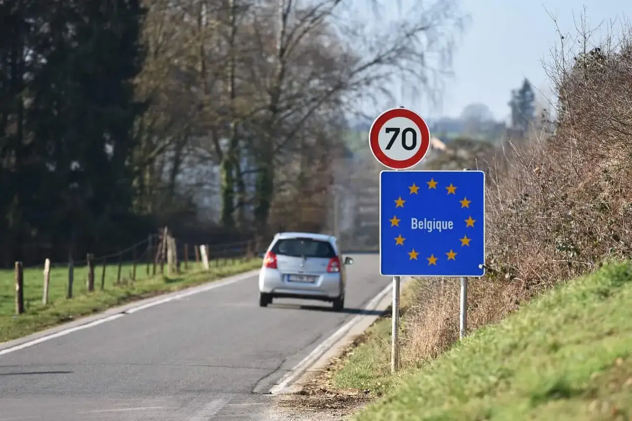 Znak graniczny Belgii, gdzie potrzebny paszport, na tle drogi i jadącego samochodu.