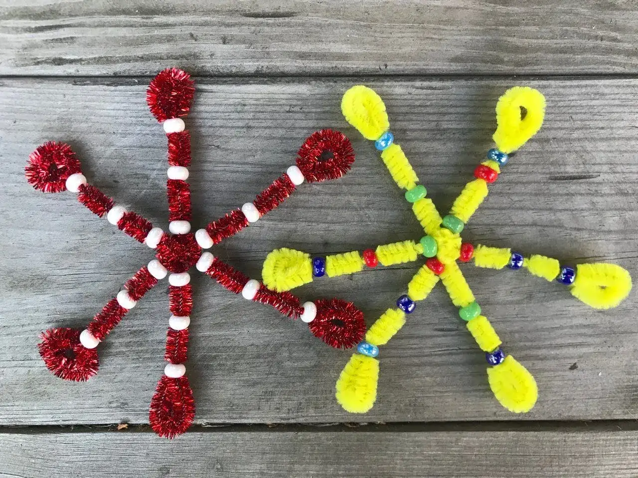 Dwie ręcznie robione gwiazdki z koralików i drucików kreatywnych: jedna czerwona z białymi koralikami, druga żółta z kolorowymi koralikami.