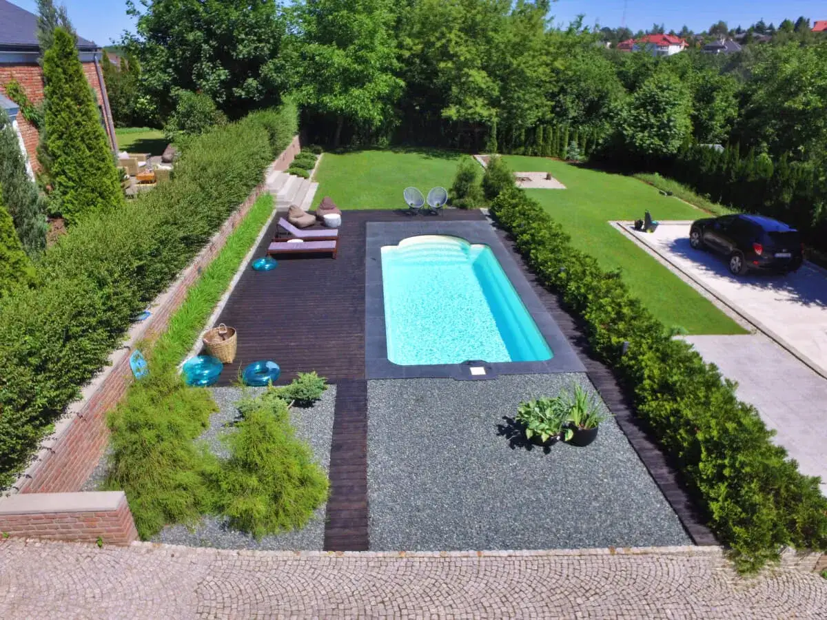 Nowoczesny basen w ogrodzie inspiracje: czysty błękit wody, drewniany taras, leżaki i bujna zieleń tworzą oazę spokoju.