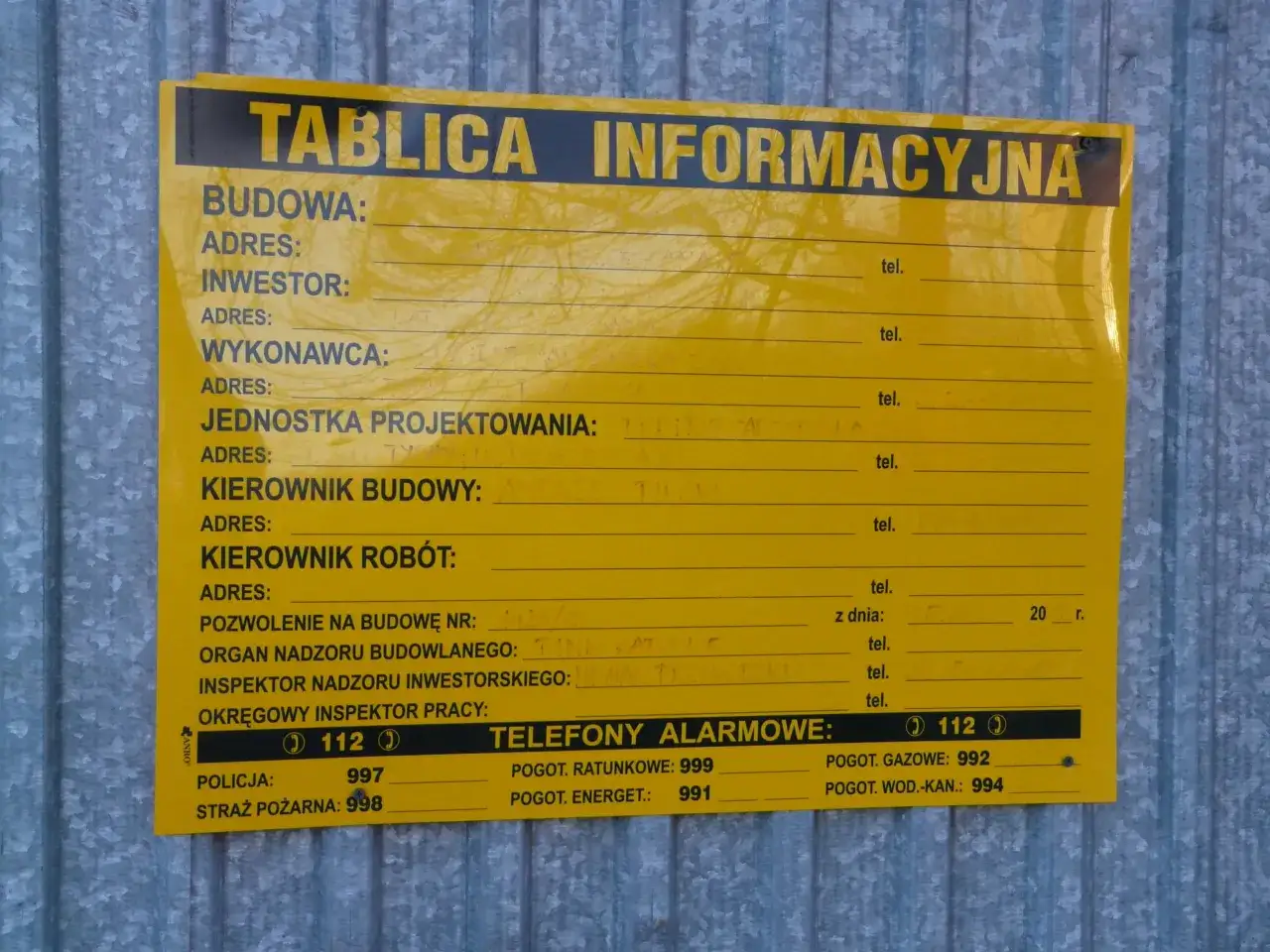 Tablica informacyjna na budowie. Widoczne pola na dane inwestora, kierownika budowy, numer pozwolenia na budowę i telefony alarmowe. Pokazuje, ile jest ważne pozwolenie na budowę.