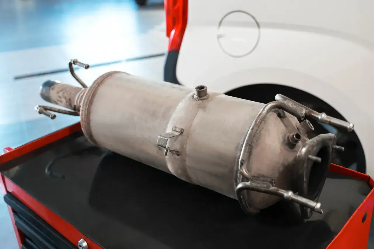Filtr DPF leżący na czerwonej skrzynce narzędziowej, obok białego samochodu.