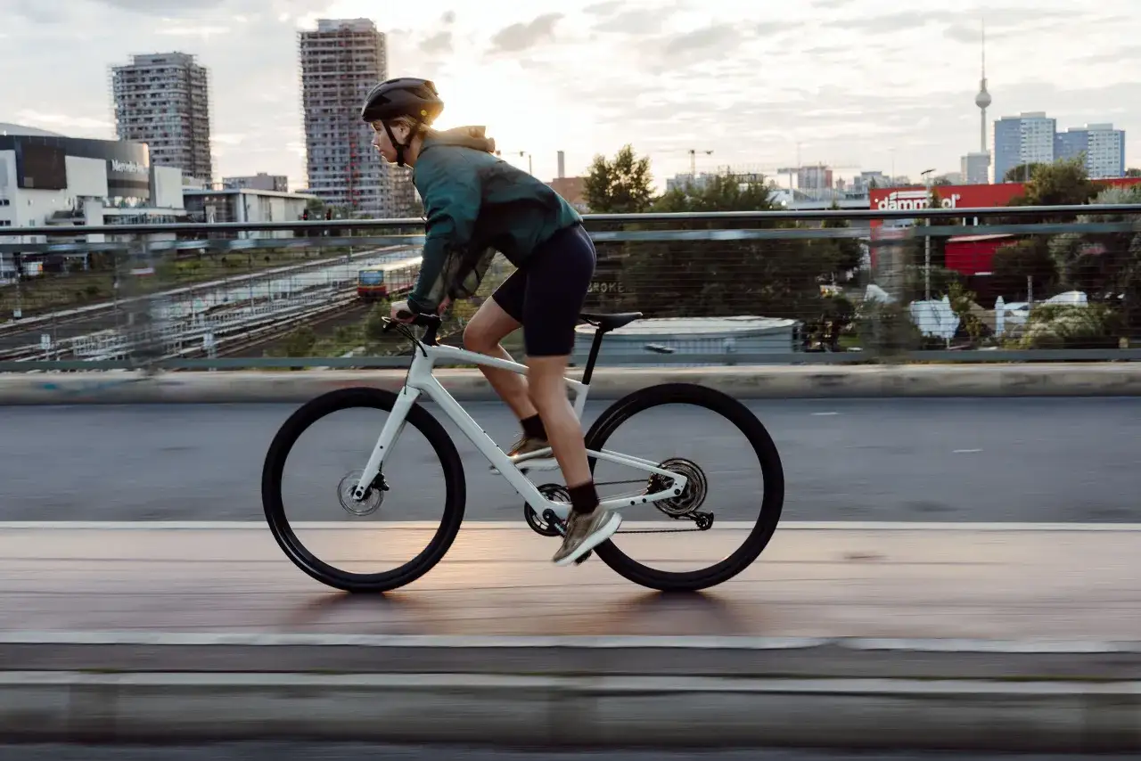 Kobieta w kasku i sportowym stroju jedzie na białym rowerze crossowym po moście, z miejskim krajobrazem w tle.