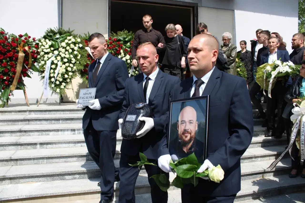 Mężczyźni w garniturach niosą urnę, tabliczkę z imieniem i zdjęcie zmarłego, otoczeni wieńcami pogrzebowymi.