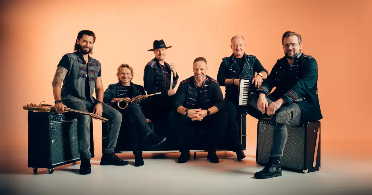 Sechs Männer einer Band posieren vor orangefarbenem Hintergrund, einige sitzen, einer steht.
