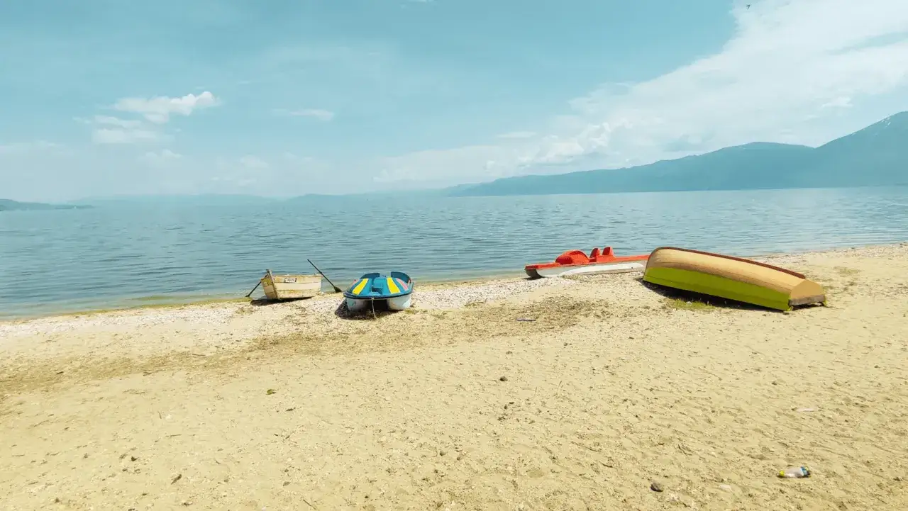 Spokojny brzeg Jeziora Ochrydzkiego w Albanii. Kilka łódek, w tym kolorowa łódka i czerwona rowerowa, odpoczywa na piaszczystej plaży.