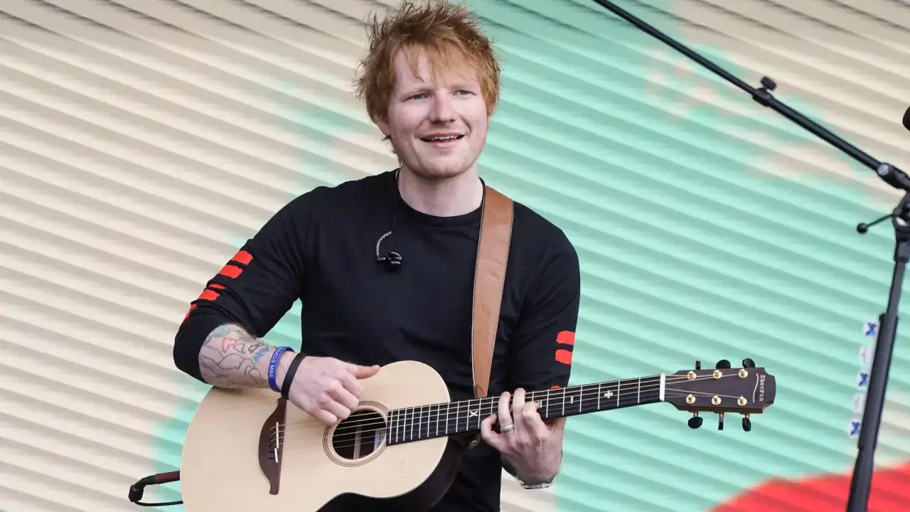 Ed Sheeran gra na gitarze akustycznej na tle pofalowanej, kolorowej kurtyny.
