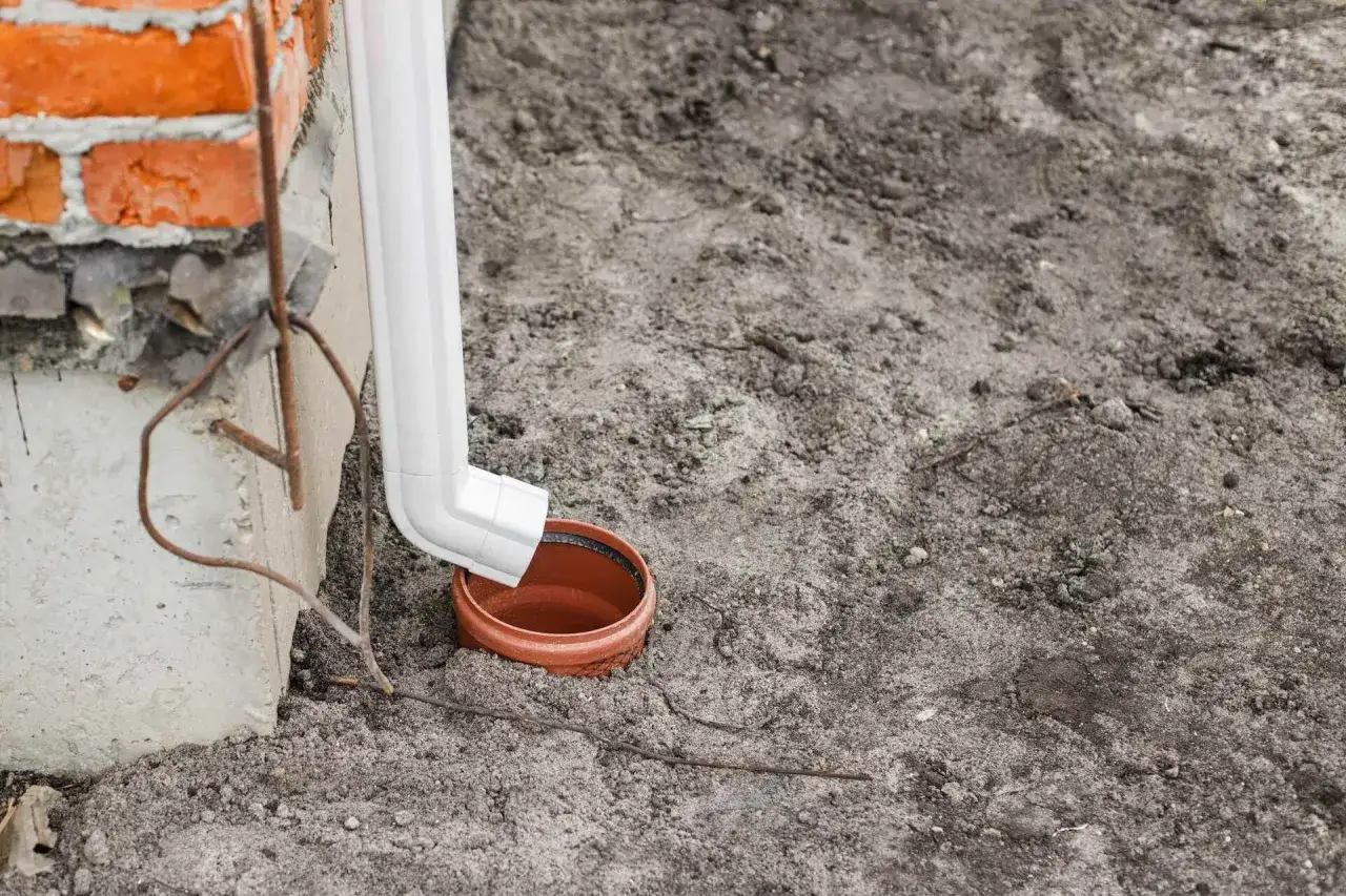 Biała rura spustowa odprowadza wodę do brązowej studzienki na ziemi.