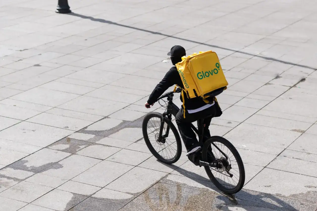 Kurier Glovo na rowerze z żółtą torbą dostawczą jedzie po chodniku.