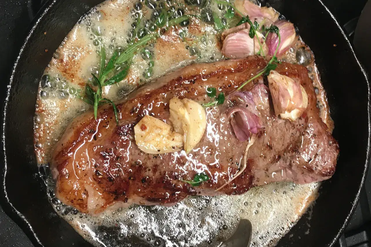 Soczysty stek z rostbefu skwierczy na maśle z czosnkiem i rozmarynem na patelni.