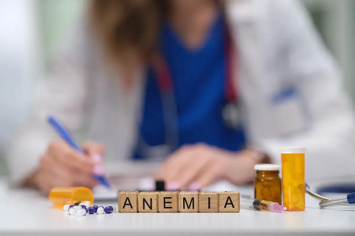 Lekarz zapisuje zalecenia dotyczące leczenia anemii u starszej osoby. Na stole leki i kostki z napisem ANEMIA.