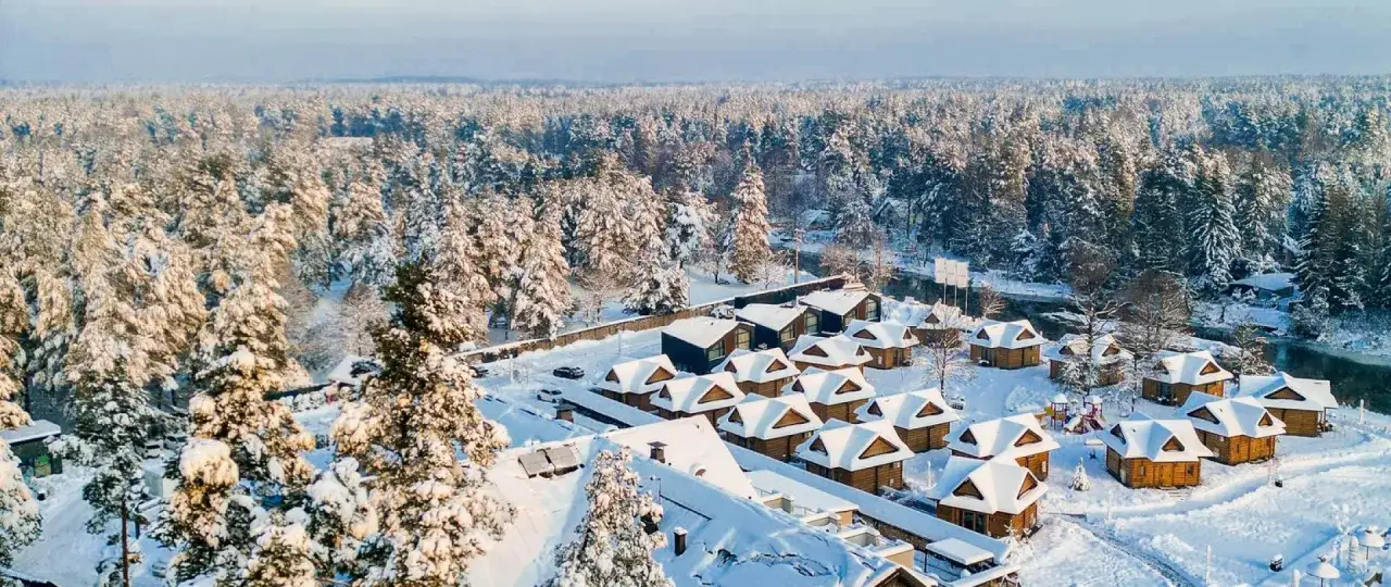 Zimowy krajobraz z lasem i śniegiem. W dole widać kompleks drewnianych domków, przypominających bacówkę na Bundówkach, otoczony ośnieżonymi drzewami.