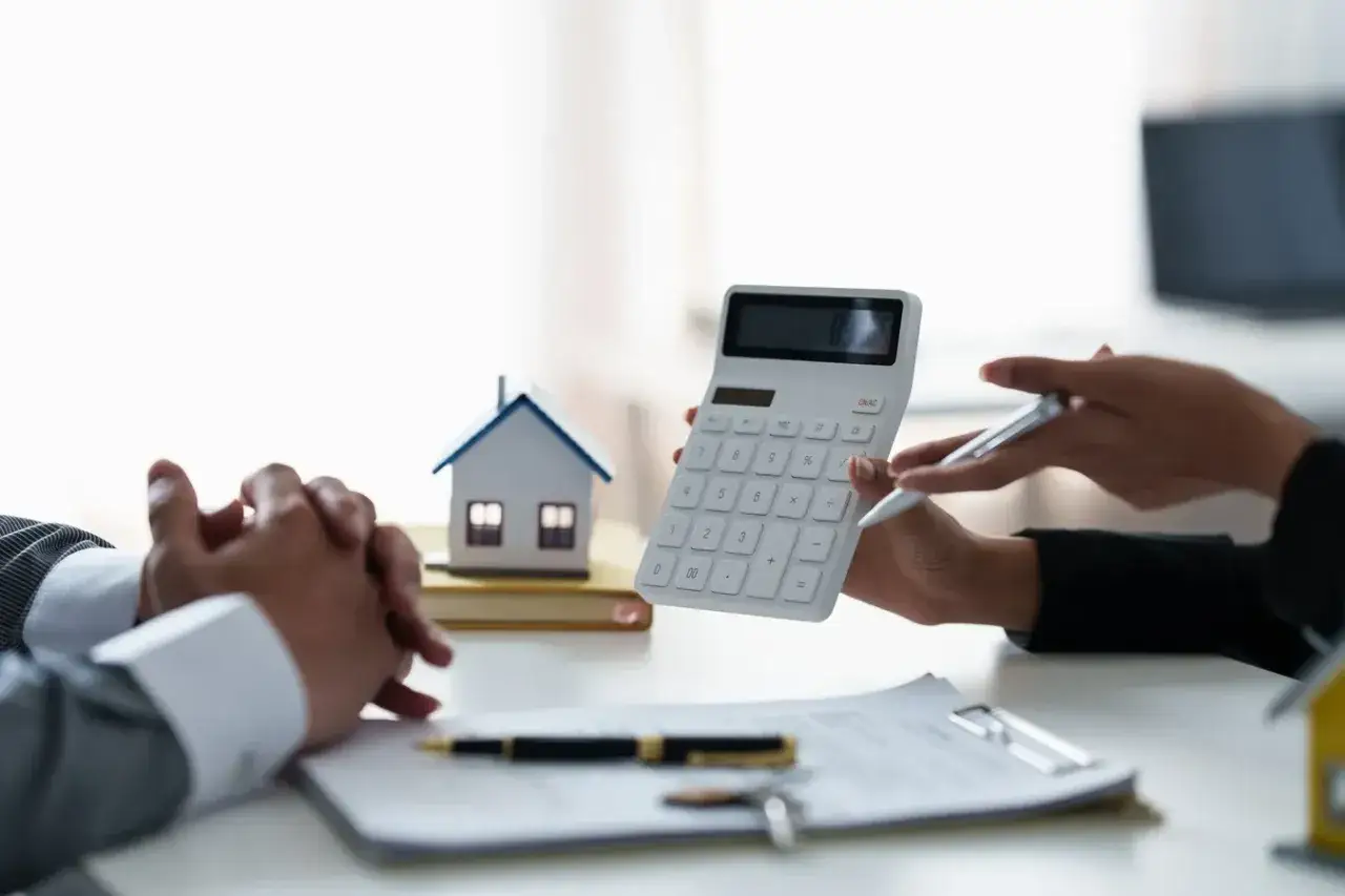 Dwie osoby przy biurku z kalkulatorem i modelem domu. Rozmowa o tym, ile zarabia notariusz miesięcznie.