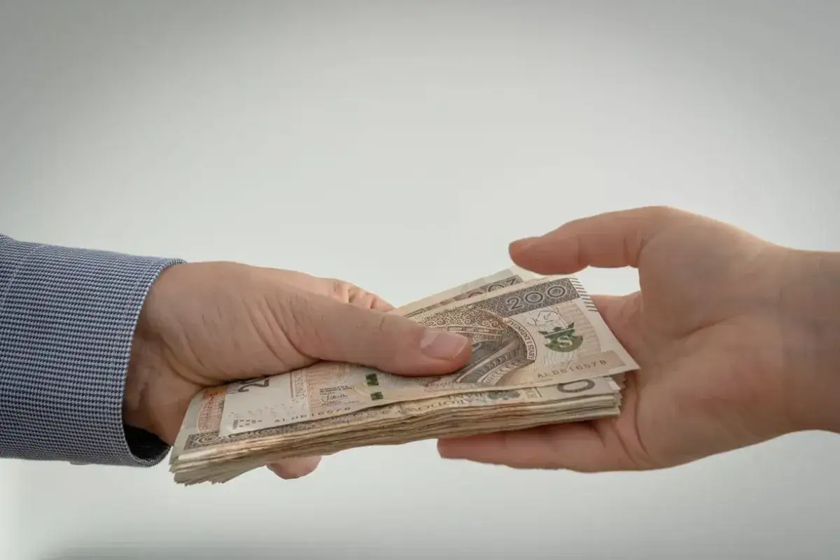 Dwie dłonie przekazujące sobie stos banknotów 200 zł.