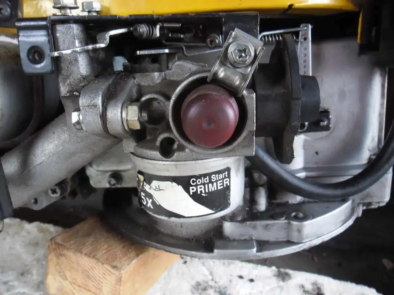 Zbliżenie na gaźnik kosiarki spalinowej z czerwonym przyciskiem "Cold Start PRIMER". Widoczne elementy mechanizmu, które mogą być pomocne, gdy zastanawiasz się, jak ustawić obroty w kosiarce spalinowej.