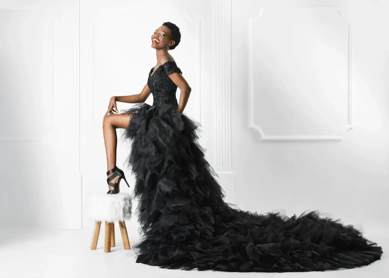 Uśmiechnięta kobieta w ekstrawaganckiej czarnej sukni ślubnej z falbanami, siedząca na puszystym stołku.