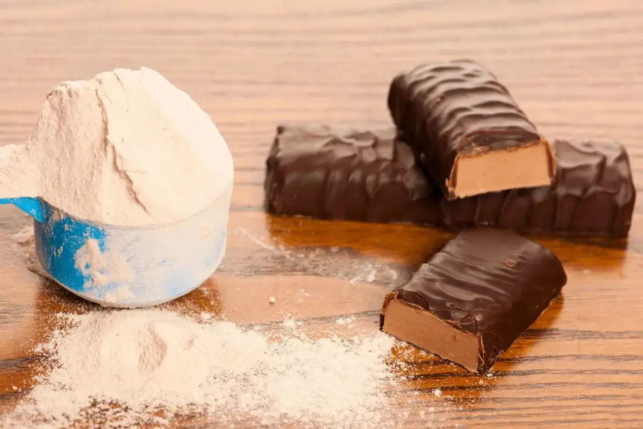 Miarka z białym proszkiem i czekoladowe batony proteinowe. Czy batony proteinowe tuczą? Zależy od składu i ilości.