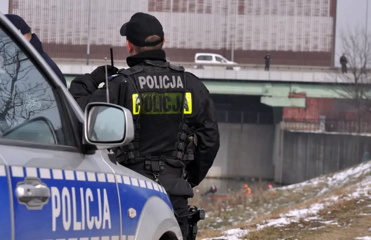 Policjant w kamizelce z napisem "POLICJA", zgodnej z rozporządzeniem w sprawie umundurowania policjantów, stoi obok radiowozu.