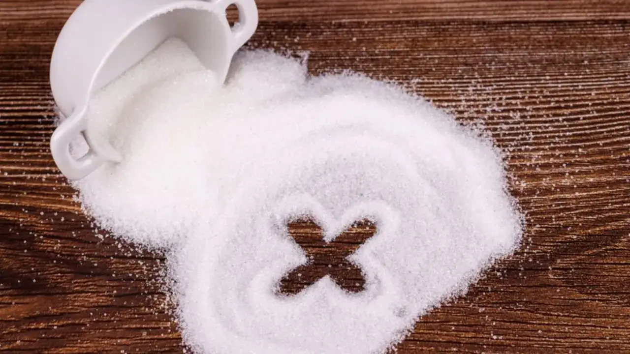 Biały cukier rozsypany z filiżanki na drewnianym blacie. W cukrze wyrysowano znak "X", symbolizujący ograniczenie spożycia. Zastanawiasz się, ile cukru dziennie jest dla Ciebie bezpieczne?
