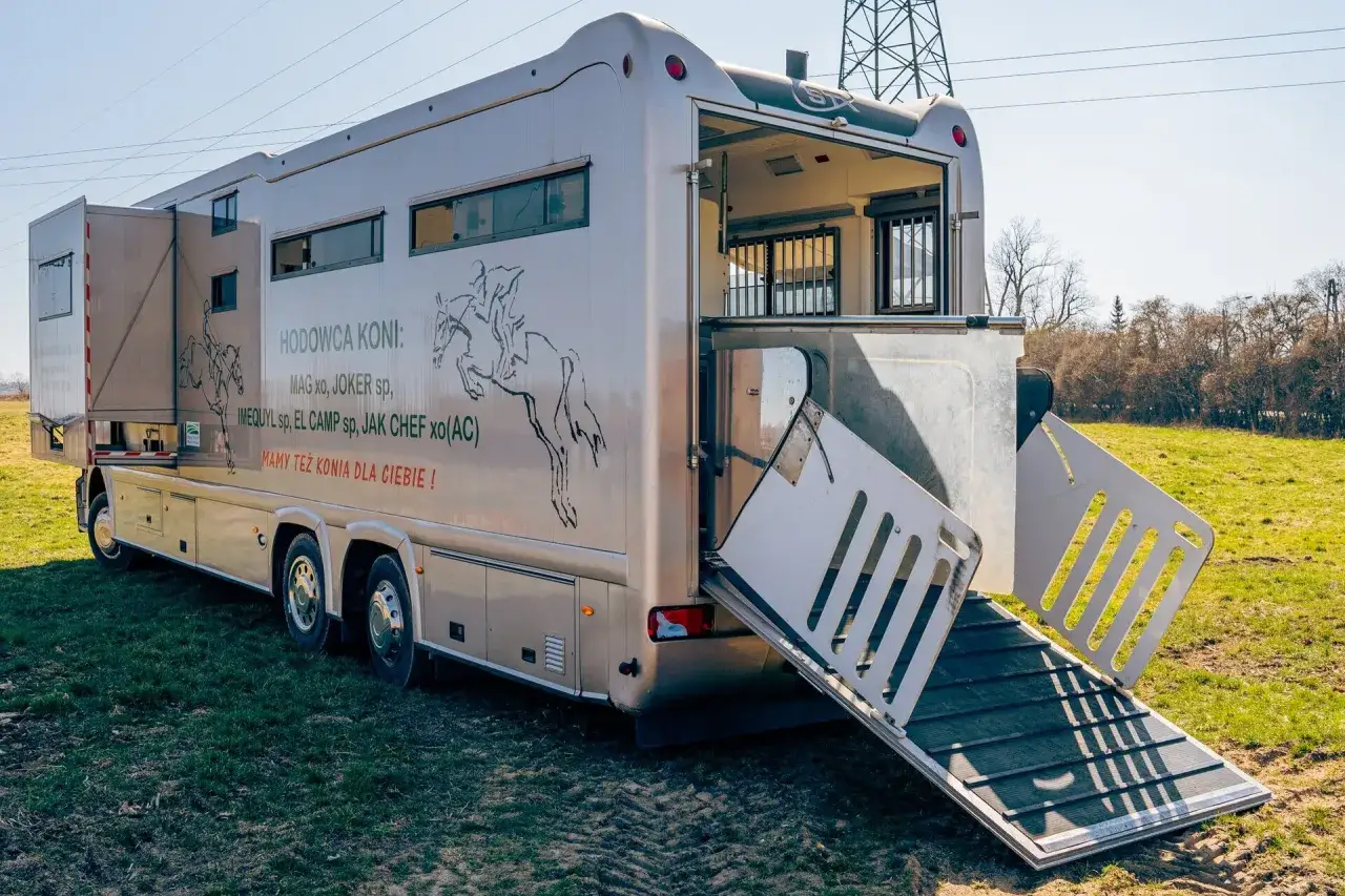 Duża przyczepa na konie z otwartą rampą, gotowa do załadunku. Na boku widnieje napis "Hodowca Koni" i rysunki jeźdźców.
