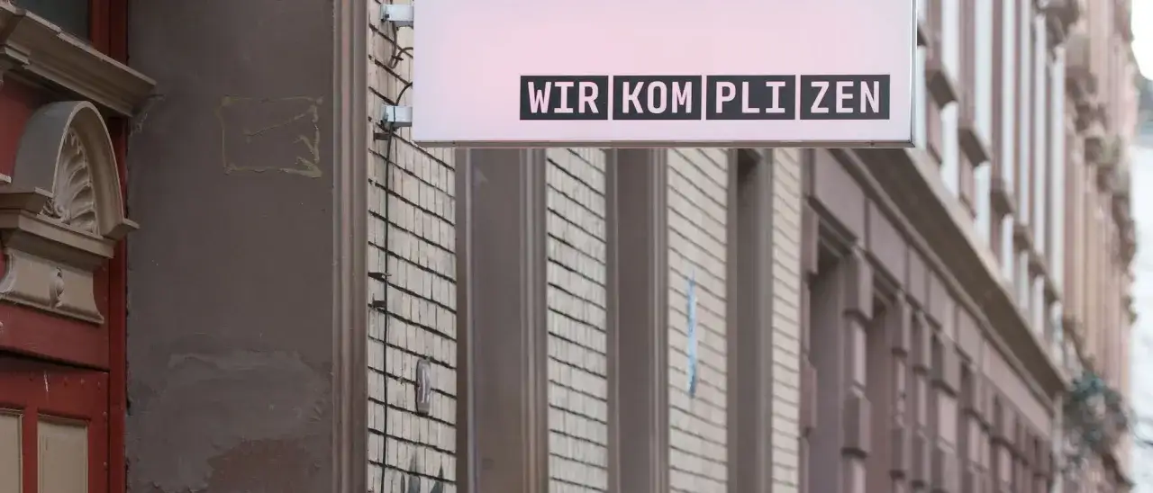 Leuchtreklame mit dem Schriftzug "WIR KOMPLIZEN" über einer Fassade.