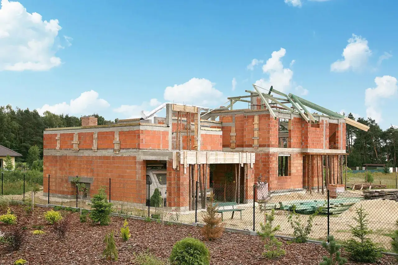 Budowa domu z cegły, widoczne zbrojenie stropu i ścian. W tle las, niebo z chmurami.