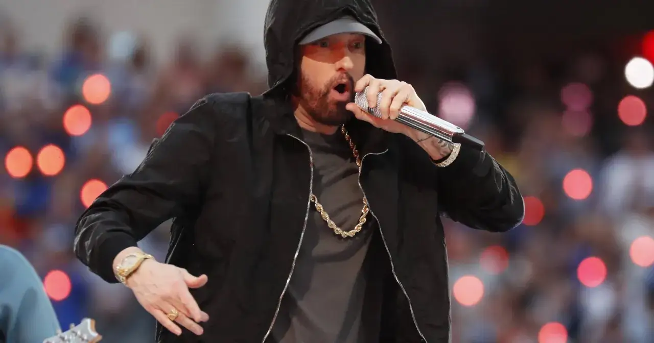 Eminem w czarnej bluzie z kapturem śpiewa do mikrofonu, w złotym łańcuchu i zegarku.