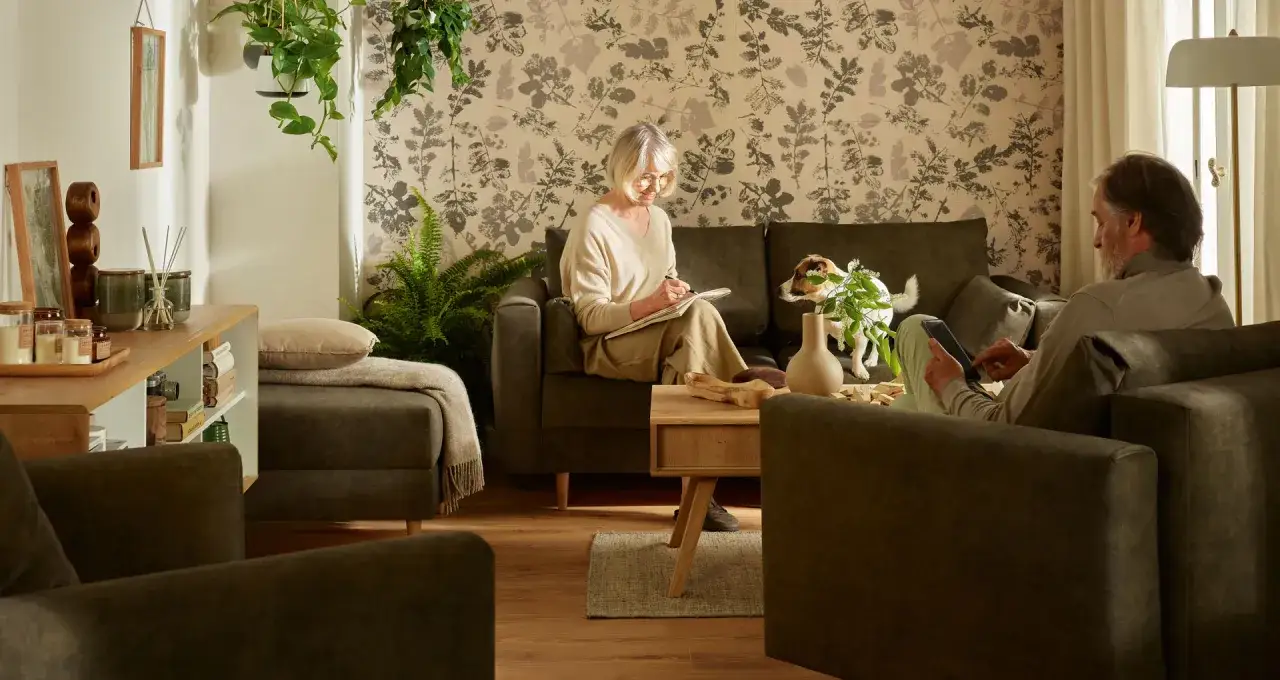 Przytulny salon, gdzie kobieta pisze, a mężczyzna korzysta z tabletu. Idealne inspiracje, jak urządzić pokój dla seniora, z wygodnymi meblami i roślinnością.