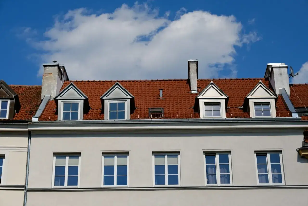 Czerwony dach z lukarnami i kominami nad jasnoszarą elewacją.
