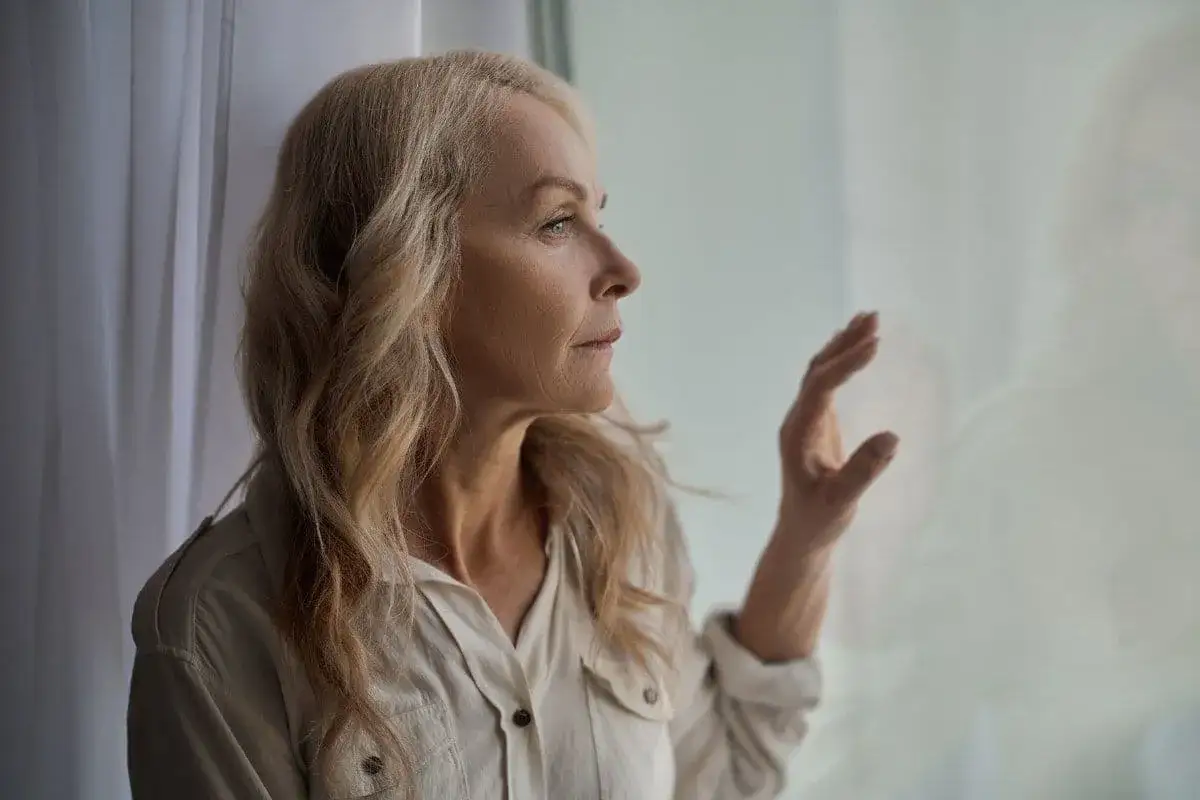 Starsza kobieta z blond włosami patrzy przez okno, zamyślona.