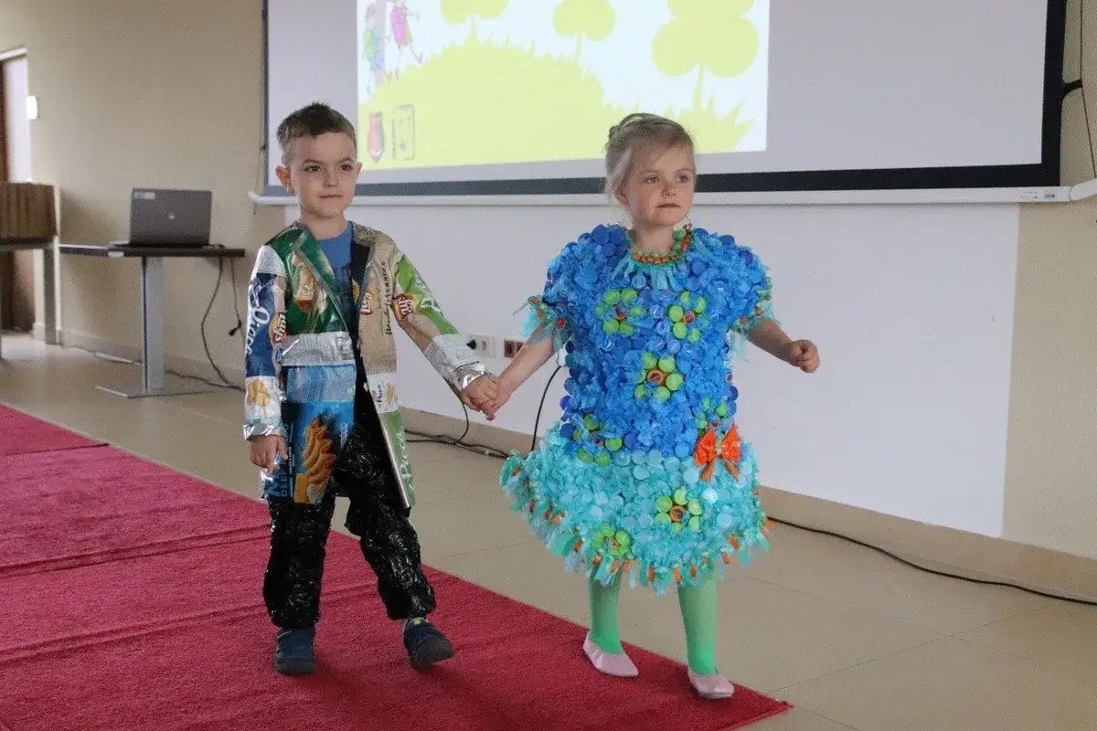 Dzieci prezentują stroje ekologiczne. Dziewczynka w niebieskiej sukience z nakrętek, chłopiec w kurtce z opakowań. Inspiracja, jak zrobić strój ekologiczny dla dziewczynki.