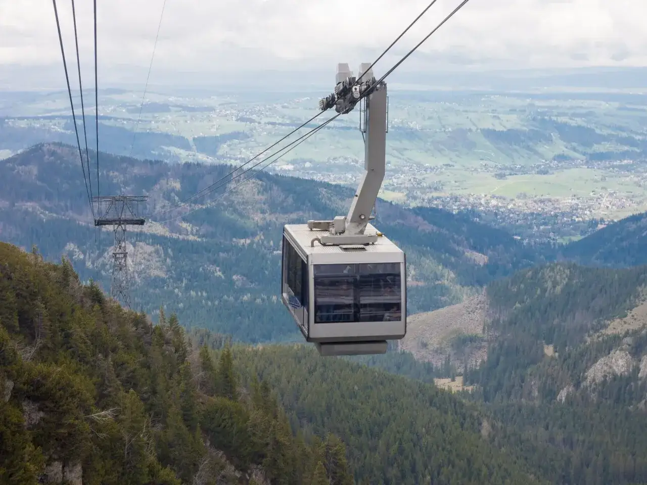 Kolejka gondolowa nad lasem i doliną z widocznym miastem w tle.