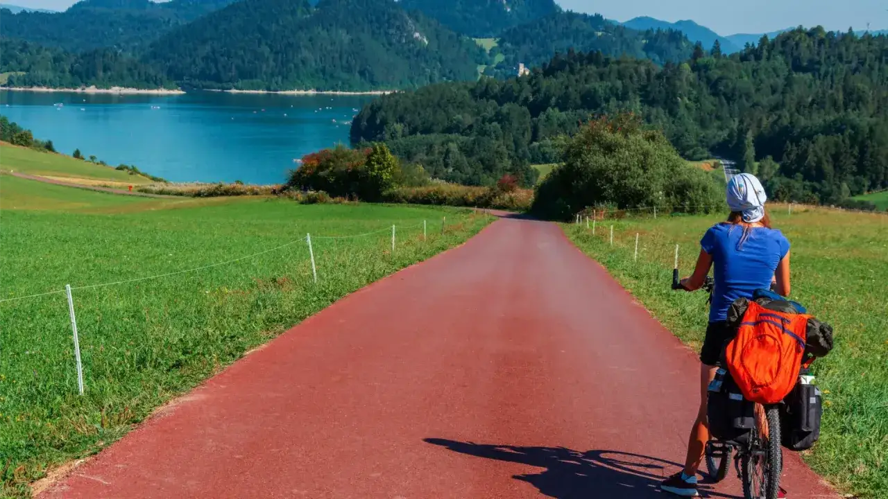 Kobieta na rowerze z bagażem na czerwonej ścieżce rowerowej. Piękna trasa Velo Dunajec z widokiem na jezioro i góry.