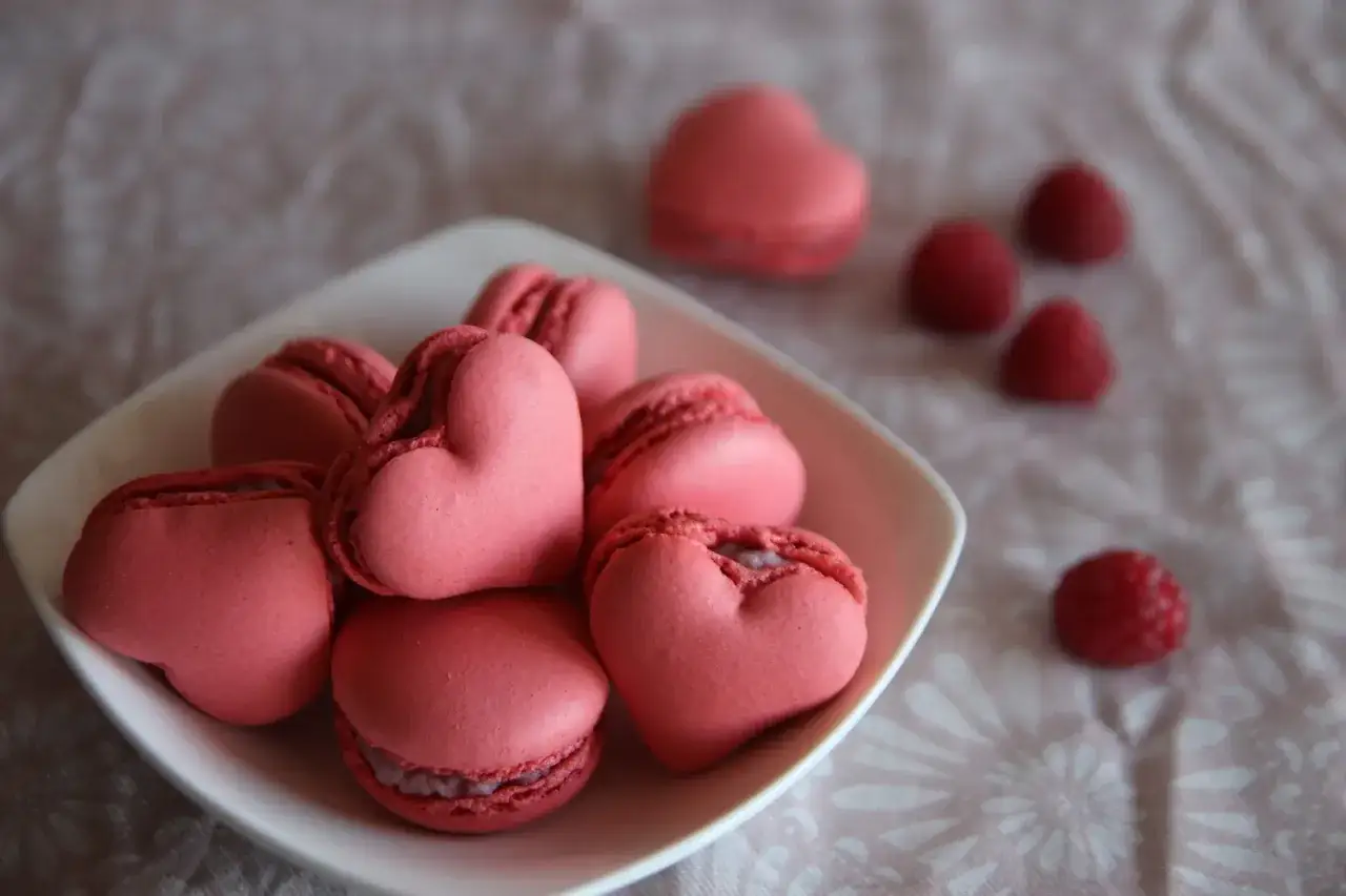 Różowe makaroniki w kształcie serc na talerzyku, obok maliny. Idealne na Walentynki, oto prosty makaroniki przepis.