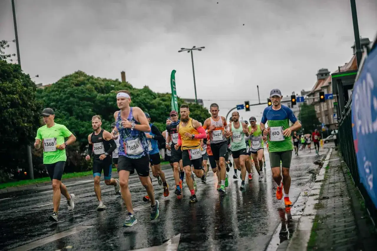 Zawodnicy biegną w deszczu podczas maratonu. Mokra nawierzchnia odbija światła, a biegacze w kolorowych strojach walczą o jak najlepszy czas.