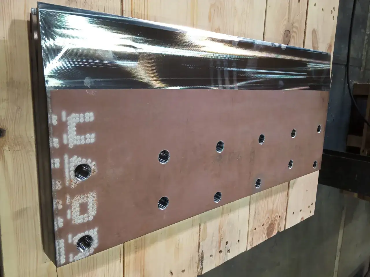 Metalowy element z otworami i oznaczeniami, leżący na drewnianych belkach.