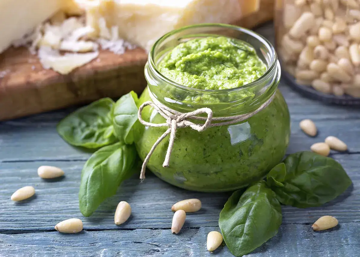 Słoik domowego pesto z bazylii, otoczony świeżymi liśćmi bazylii i orzeszkami pinii. Obok kawałki parmezanu. Idealne do makaronu.