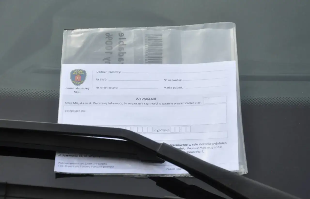 Wezwanie od Straży Miejskiej za złe parkowanie. Dokument zawiera pieczęć Straży Miejskiej Warszawa, numer alarmowy 986 i pole do wpisania przyczyny wezwania.