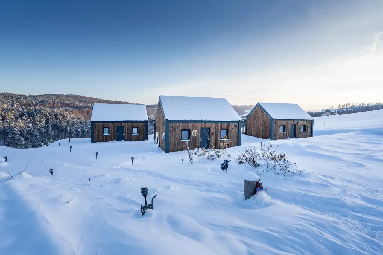 Drewniane domki na śnieżnym wzgórzu, idealne miejsca na ferie zimowe. W tle las, błękitne niebo.