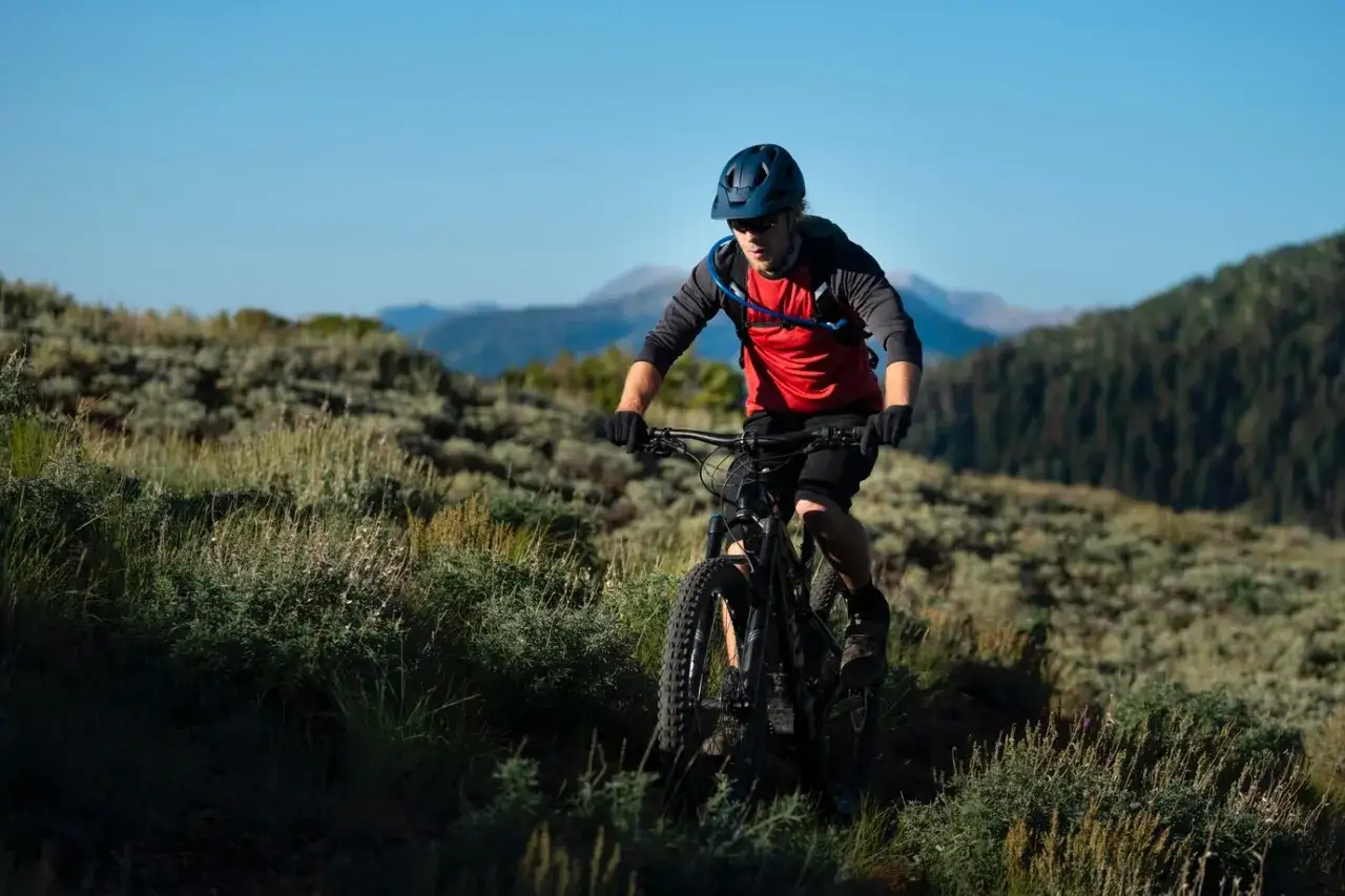 Kolarz MTB w kasku i czerwonej koszulce z plecakiem wodnym, jadący po górskim szlaku. Utrzymuje prawidłową pozycję na rowerze mtb, pochylony do przodu.
