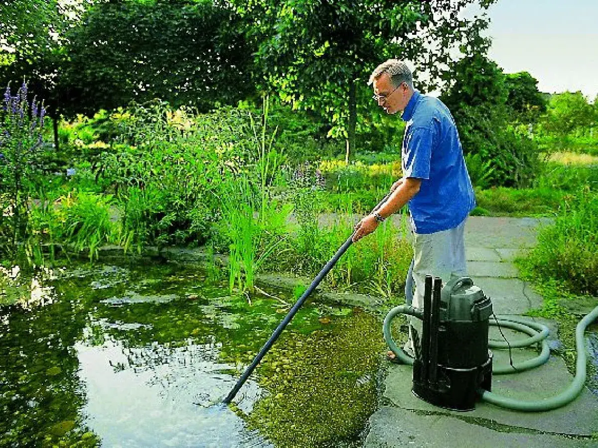 Mężczyzna wykonuje czyszczenie oczka wodnego za pomocą specjalnego odkurzacza. Wokół bujna zieleń.