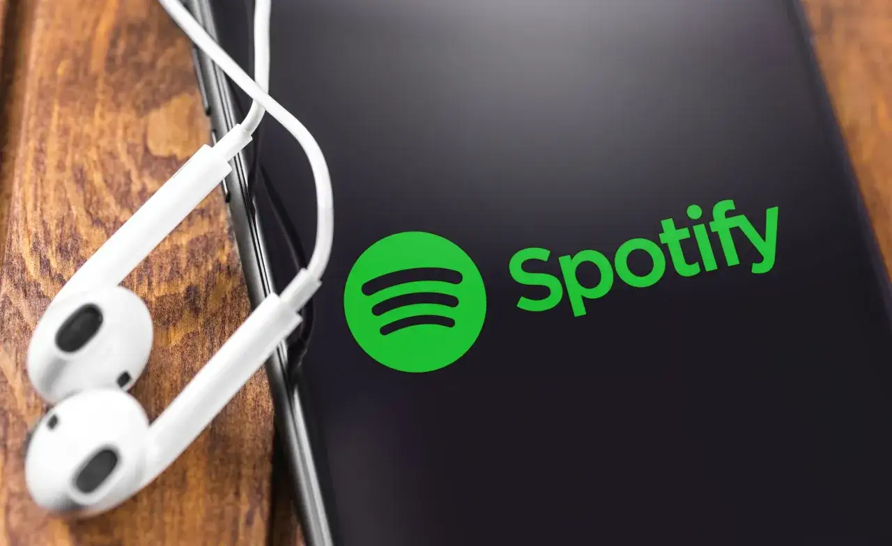 Logo Spotify na ekranie telefonu z białymi słuchawkami na drewnianym tle.