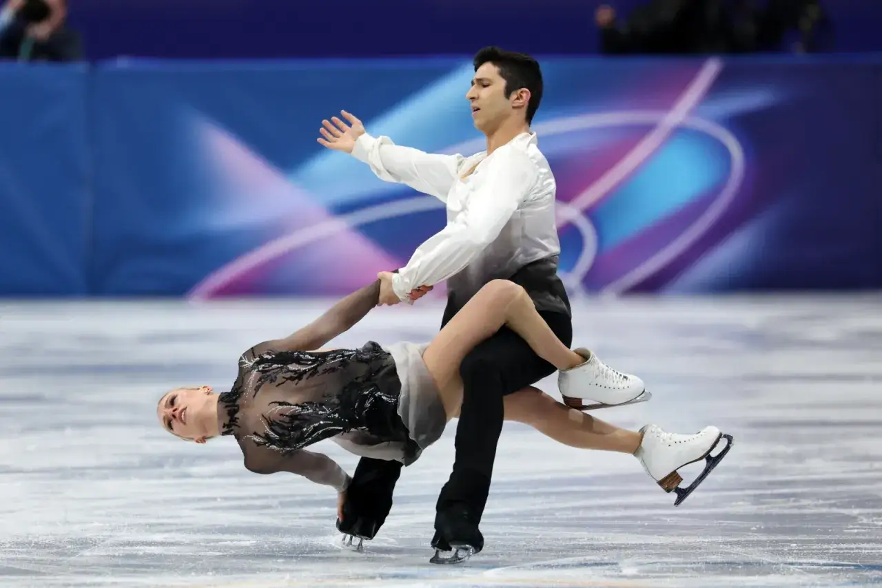 Para tańców na lodzie wykonuje skomplikowaną figurę. Kobieta w czarnej sukience leży na lodzie, podtrzymywana przez partnera w białej koszuli.