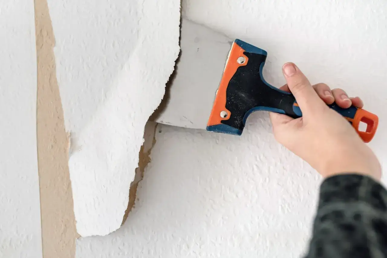 Dłoń zeskrobująca starą tapetę ze ściany za pomocą szpachelki.