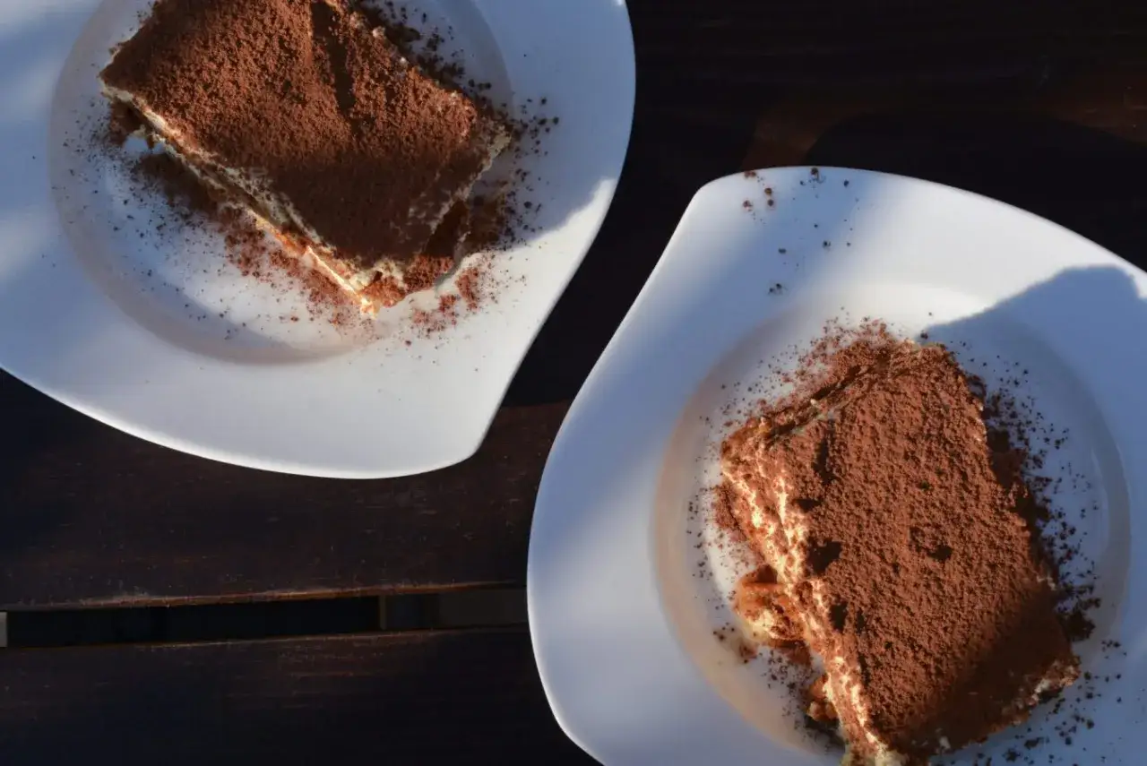 Dwa kawałki tiramisu posypane kakao na białych talerzach. Idealne do mocnej kawy, która podkreśli smak.