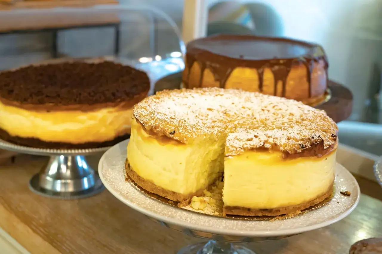 Trzy serniki: jeden z polewą czekoladową, drugi z kruszonką, a trzeci oprószony cukrem pudrem, z wyciętym kawałkiem.