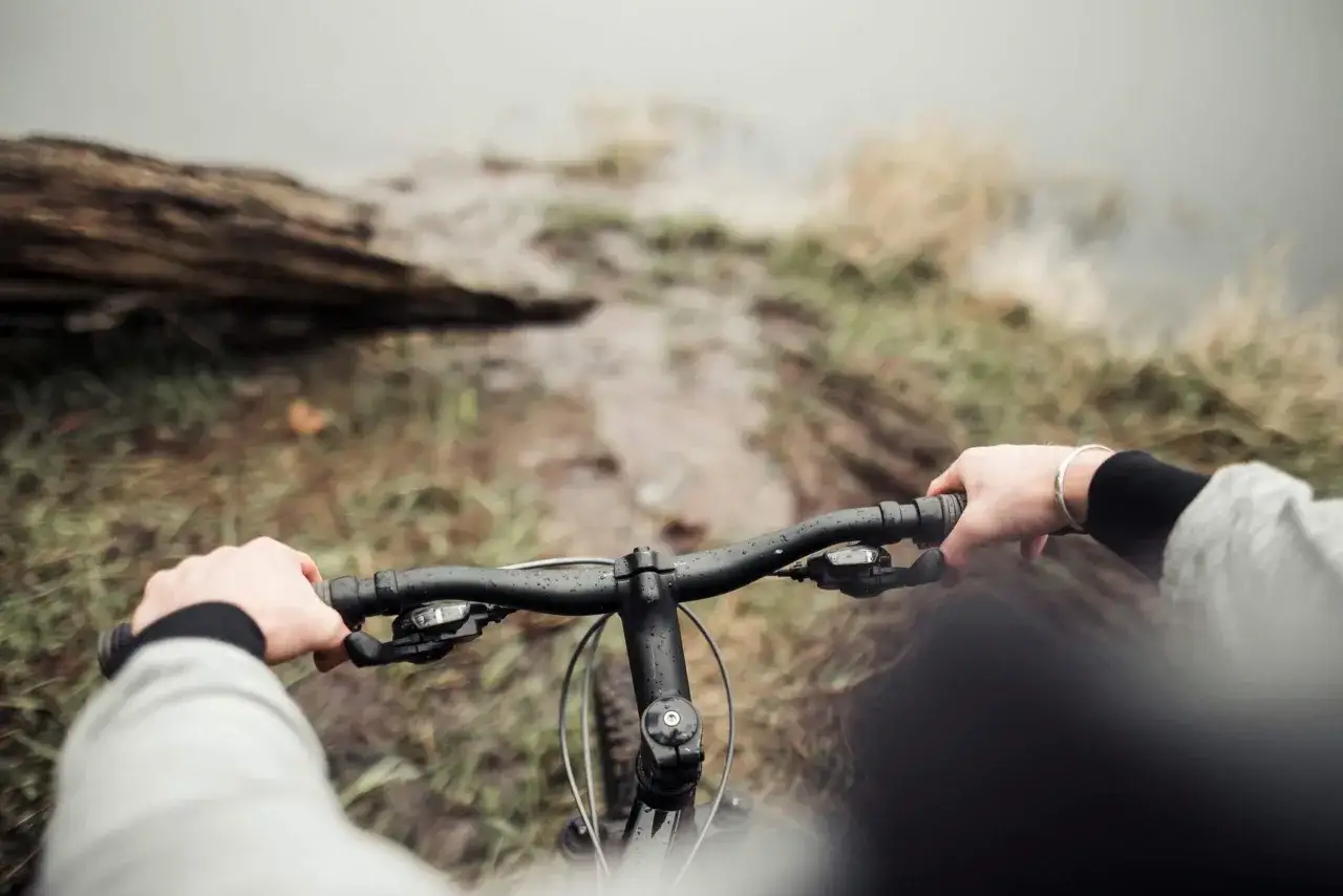 Ręce na kierownicy roweru, błotnista ścieżka i widok na wodę. To właśnie dlatego warto jeździć rowerem – bliskość natury i wolność.