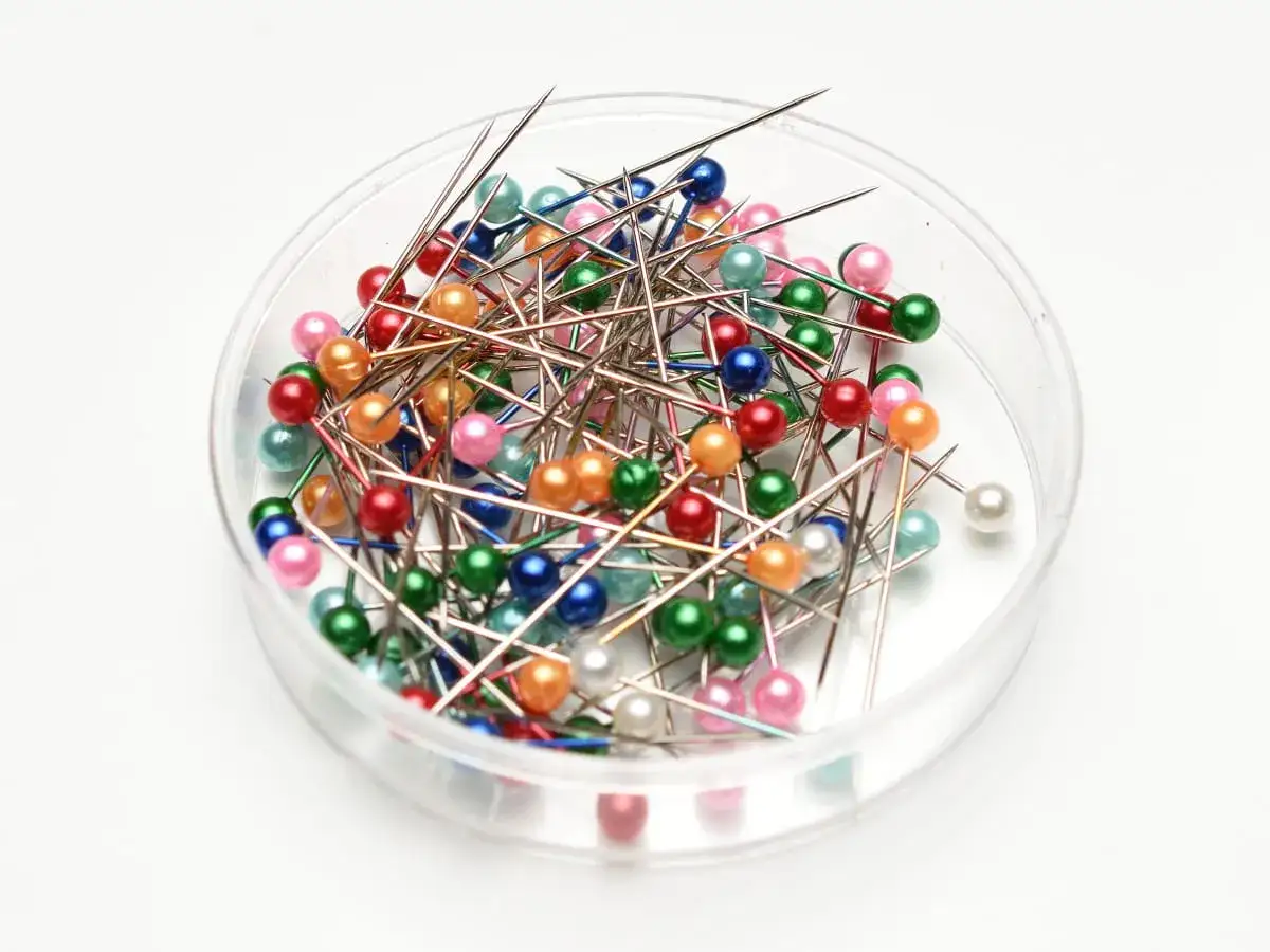 Kolorowa główka szpilki w przezroczystym pojemniku. Wiele igieł z kolorowymi, perłowymi główkami w odcieniach zieleni, różu, czerwieni i błękitu.